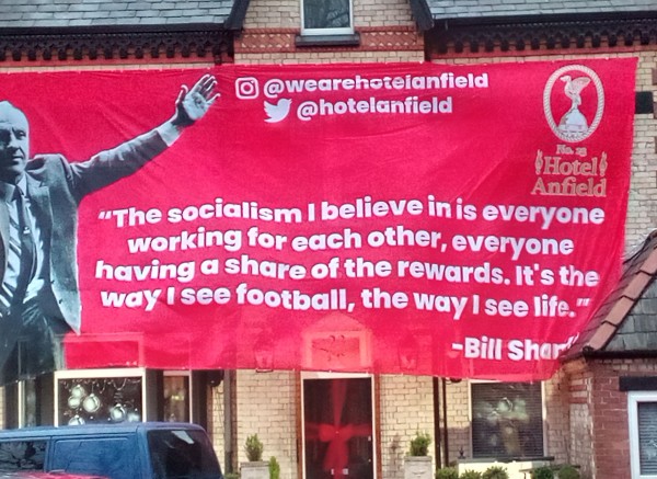 Banner in red on the Hotel Anfield showing a picture of Bill Shankly left arm aloft in greeting and the statement from him - "The socialism I believe in is everyone working for each other, everyone having a share of the rewards. It's the way I see football, the way I see life"