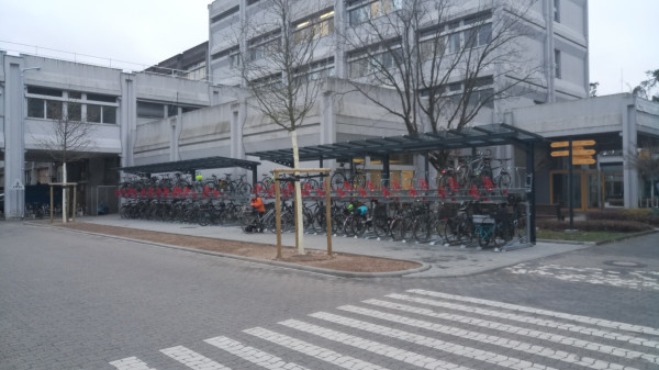 Foto der neuen Fahrradständer gegenüber den alten Felgenbrechern. Die Vorrichtung ist zweistöckig, überdacht und (nicht gut zu sehen) beleuchtet. Installiert sind etwa 80 Plätze, sie stehen mehr als halbvoll.