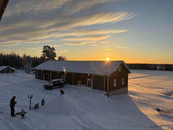 The sun is just visible above a barn and snowy fields. Three children and a man are outside in the snow.