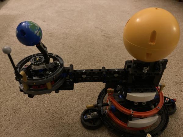 A Lego orrery of the Sun-Earth Moon system sits on a boring beige carpet. The sun is on the right. The Earth, on the left, has its northern hemisphere tilted away from the sun, reflecting its actual orientation during the Northern hemisphere winter solstice. The moon is to the left and a little in front of the Earth. 