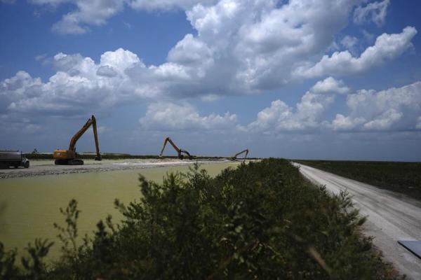 Large crawler tractor shovels at work along a gray-green stretch of water under a sunny sky with clouds. This is the U.S. Army Corps of Engineers building the Everglades Agricultural Area Reservoir, intended to store polluted water from Lake Okeechobee and agricultural runoff so it can be cleaned in an adjacent stormwater treatment area before being released to flow south into the Everglades. AP photo by Rebecca Blackwell.