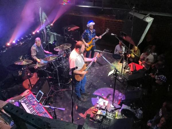 Cris Jacobs (guitar) onstage with his Very Jerry band, including a drummer, bassist, and keyboard player. The keyboard player is out of frame, but one of his keyboards, likely a Nord Electro, can be seen