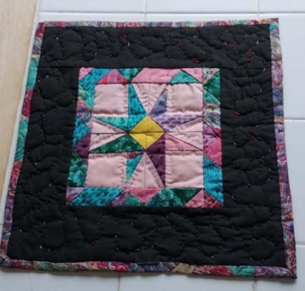 a quilt, it is a star made of yellow, blue, purple, and green triangles on a pink square with a multi-colored border, on a larger black square, a multi-colored thin border around it 