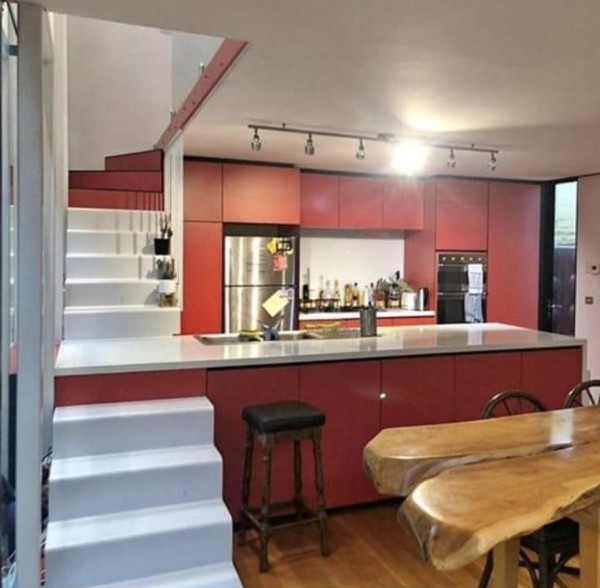 So photo of a red and white painted kitchen that has stairs integrated into the smooth countertop. You can see a fridge, a bar space, and an oven in the background lit up with track lighting. A bar stool sits next to the bottom part of the stairs, and a few plants have been placed on the stair (where you step) as decoration next to where you would stand by the kitchen sink... centimeters away.