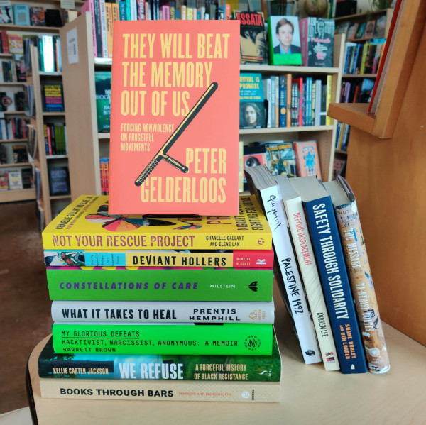 A photo of nonfiction titles from Firestorm's 2024 Bestsellers & Collective Picks list. The books are stacked on the edge of a table and bookshelves are visible in the background. At the top of the stack is a faced copy of "They Will Beat the Memory Out of Us: Forcing Nonviolence on Forgetful Movements."