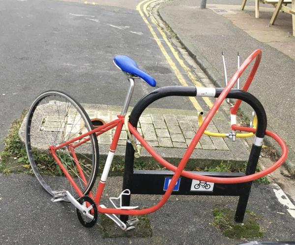 Picture shows a red racing bike bent around a Sheffield bike rack. It looks odd, and a second glance shows that the crossbar is improbably long and the knot that the whole frame is tied in is an impossibility for a bike, making it more of a surreal sculpture 