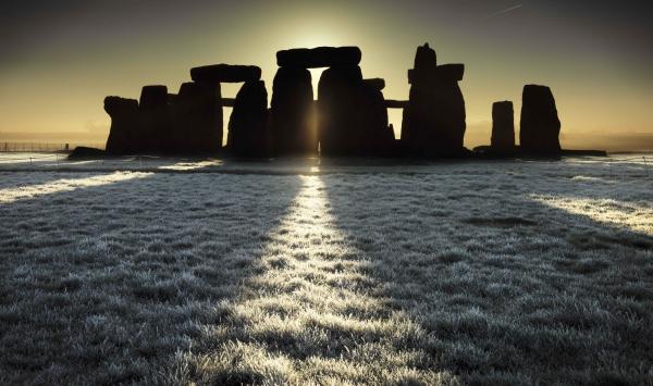 Stonehenge in winter at solstice. There’s frost on the grass, the monument is dramatically lit from behind by the winter sun low in the sky making it appear in silhouette. 