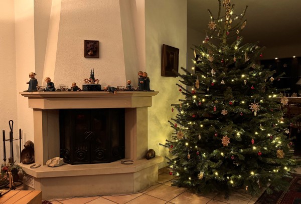 Rechts im Bild ein Weihnachtsbaum, links daneben ein offener Kamin mit geschlossenen schwarzen Türen, daneben ein Kaminbesteck, auf dem Kaminsims eine Reihe blauer Engelfiguren, dazwischen in der Mitte eine Spieluhr