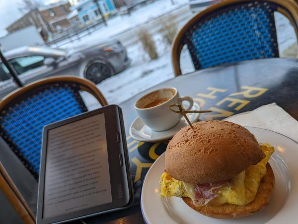 A breakfast sandwich of egg, prosciutto, and cheese alongside a cappuccino and Kobo e-reader on an outdoor cafe table; in the background is a snowy sidewalk, grass, and buildings.