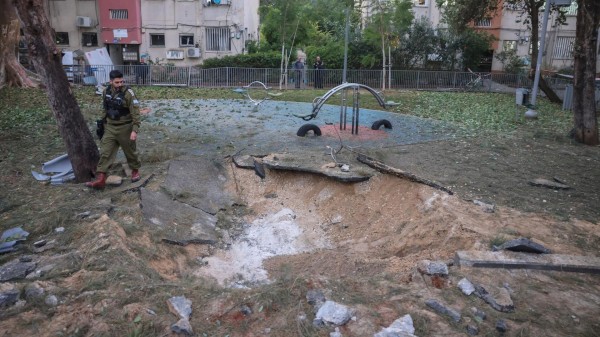 Ein Krater auf einem Kinderspielplatz. Daneben steht ein Soldat und betrachtet die Trümmer