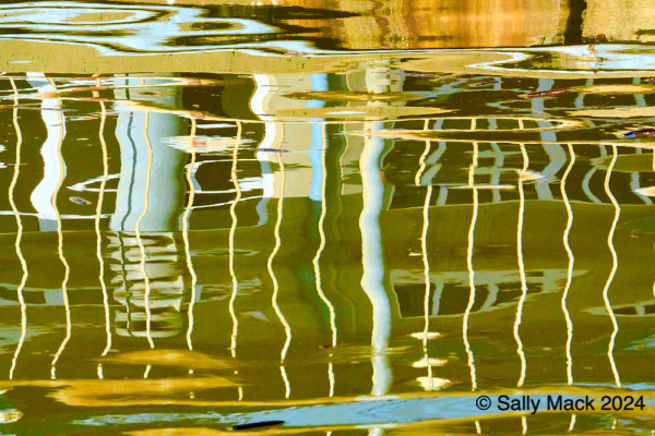 Color photo of reflections of a railing in moving, greenish water resulting in mostly vertical forms of mostly yellow and light blue. All forms are squiggily.