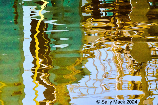 Color photo of reflections of ferry dock in moving, blue water resulting in mostly vertical forms of many colors, turquoise, yellow, pink, white, brown. All forms are squiggily.