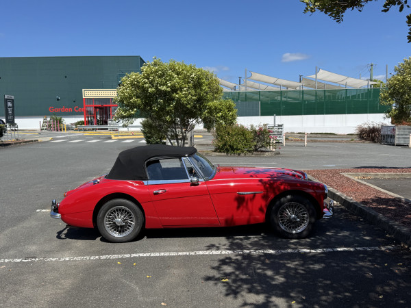 A red convertible Austin-Healey 3000 with the roof up parked in a car park. 