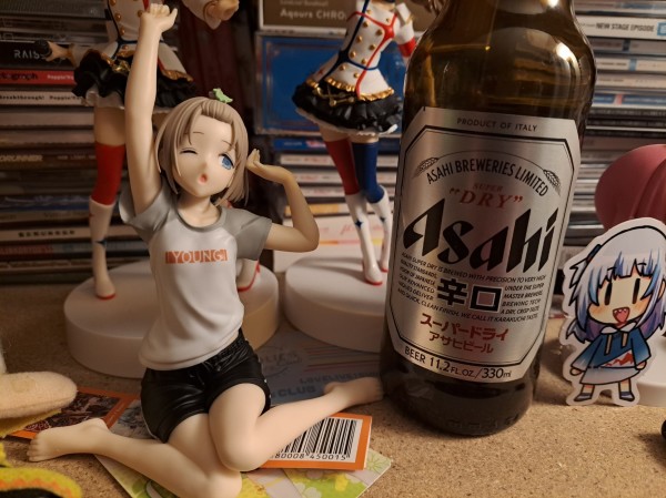 A figure of Asahi Serizawa beside a bottle of Asahi beer. Asahi has her front bangs pulled back with a dinosaur hair pin. She's wearing a white t-shirt with gray short sleeves and color. In the middle of the shirt, the word Young is written. She is also wearing black gym shorts. She is stretching as if she's waking up from bed. One eye is closed, but the eye that is open appears to be looking at the bottle of Asahi beer.