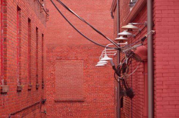 A photo I took from inside a building depicting the second story of an alley of bright red bricks. The window a the end of the alley has been bricked up, as have other windows leading down the alley. There are also power lines leading down from outside the frame to the right side of the alley where there is a line of lamps leading all the way to the dead end. A brownish gutter pipe leads down the right side as well. 