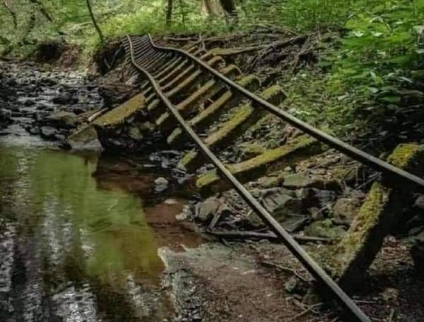 A railroad track, dilapidated, broken, missing sleeper hangs askew, the bank beneath it having collapsed into the stream running beside it