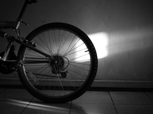 Black and white photo of a square of bright sunlight falling on a textured and also more diffusely lit wall, in front of which is the rear wheel and part of the frame of a bicycle.