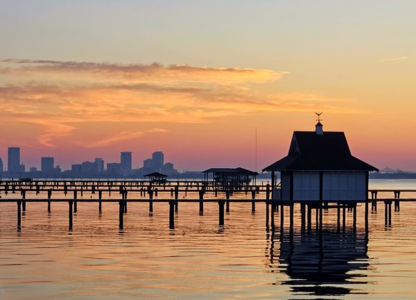 Sherbert colored sky above the silhouettes of a downtown skyline in the distance with golden reflections upon a quiet river in the foreground, where several long wooden piers reach out into the water, with covered boat docks at the end of each, resembling small homes.