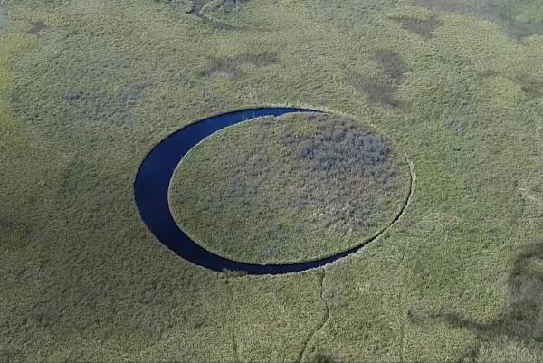 Das Bild, welches aus großer Höhe aufgenommen wurde, zeigt eine grüne Landschaft mit einem runden See in der Mitte. Im See ist eine runde Insel zu sehen. Das Ganze erinnert an ein Auge